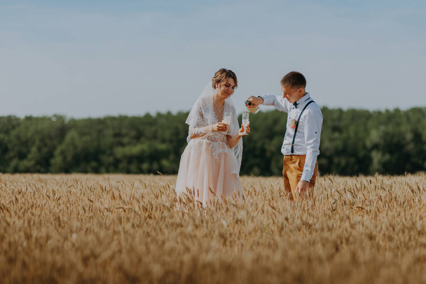 Bride and groom holding wedding champagne glasses on the background of wheat field. Happy wedding couple in wheat field. Beautiful bride in white dress and groom having fun on summer day. Just married - Fotó, kép
