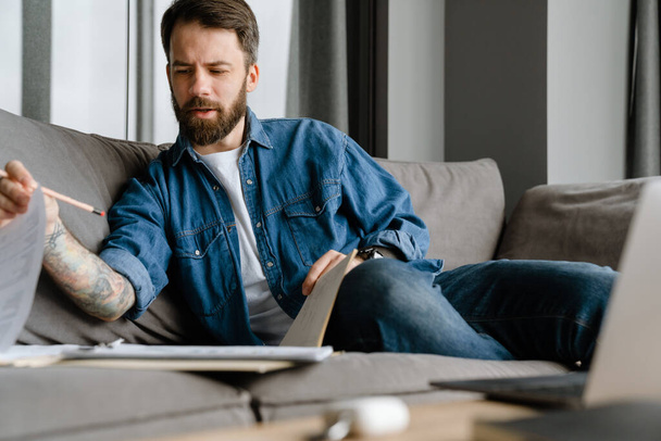 Bearded european man working with papers and laptop while sitting on sofa at home - Foto, Imagem