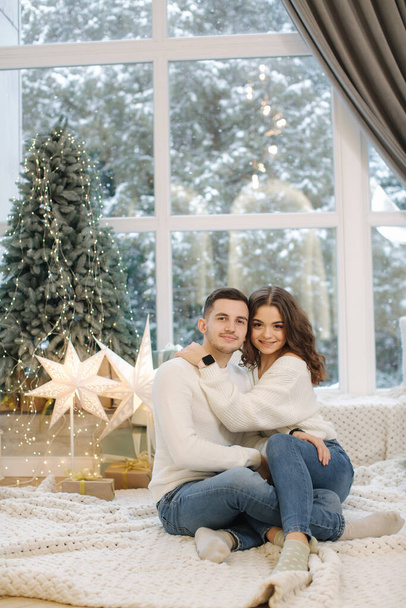 Un chico guapo con su hermosa novia se abrazan en casa en la víspera de Navidad. Ajuste árbol y nieve outsif. Humor de Navidad - Foto, imagen
