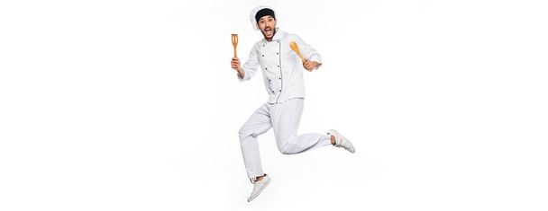 chef excitado en sombrero y uniforme saltando y sosteniendo espátula de madera y cuchara aislado en blanco, bandera - Foto, imagen