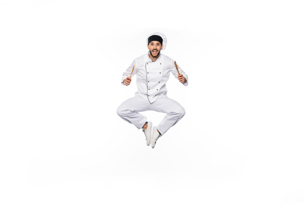 Chef alegre en sombrero y uniforme saltando y sosteniendo espátula de madera y cuchara aislada en blanco  - Foto, imagen