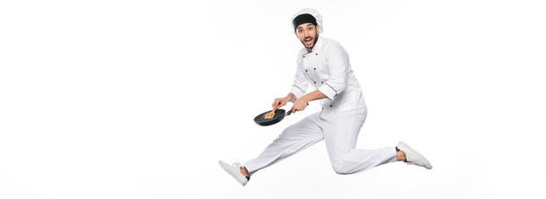 veselý kuchař v klobouku a uniformě skákání a držení pánev s dřevěnou špachtlí izolované na bílém, banner - Fotografie, Obrázek