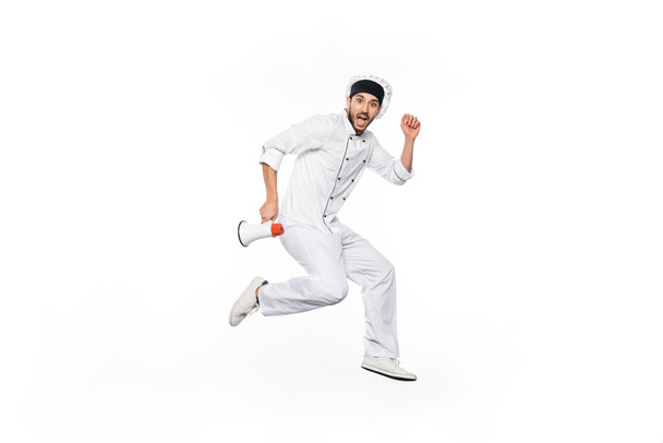 shocked young chef in hat and uniform levitating and holding megaphone isolated on white  - Фото, зображення