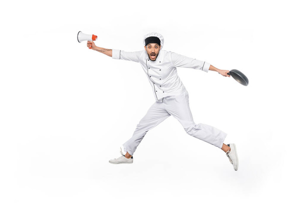 surprised young chef in hat and uniform levitating while holding frying pan and megaphone isolated on white  - Fotoğraf, Görsel