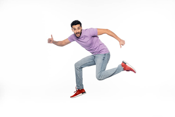 amazed man in jeans and purple t-shirt levitating while showing thumbs up isolated on white  - Fotó, kép