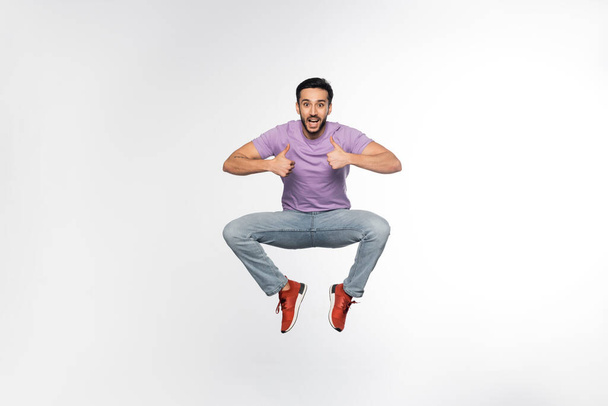 hombre sorprendido en jeans y camiseta púrpura levitando mientras muestra los pulgares hacia arriba en blanco  - Foto, Imagen