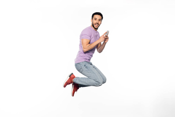 úžasný muž v džínách a fialovém tričku levitující při držení smartphonu na bílém  - Fotografie, Obrázek