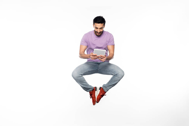 homme heureux en jeans et t-shirt violet lévitant tout en utilisant une tablette numérique sur blanc  - Photo, image