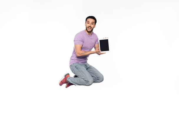 hombre sorprendido en jeans y camiseta púrpura levitando mientras sostiene la tableta digital en blanco  - Foto, imagen