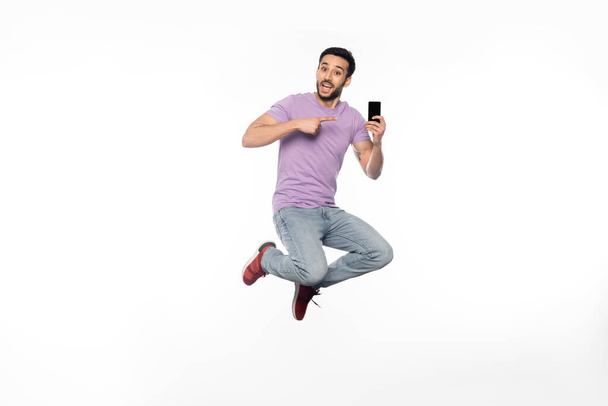 homem surpreso em jeans e camiseta roxa levitando enquanto apontava para o smartphone em branco  - Foto, Imagem