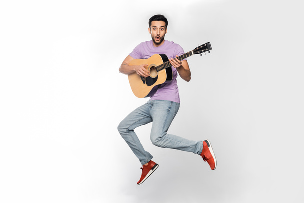 шокированный мужчина в джинсах и фиолетовой футболке, левитирующий во время игры на акустической гитаре на белом  - Фото, изображение