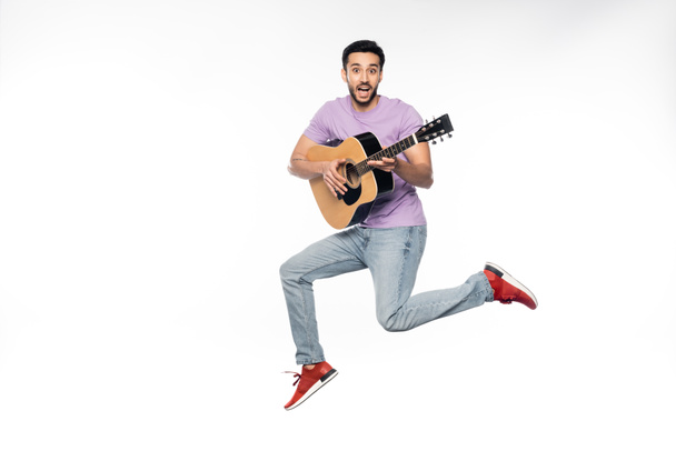 amazed man in blue jeans and purple t-shirt levitating while playing acoustic guitar on white  - Фото, зображення