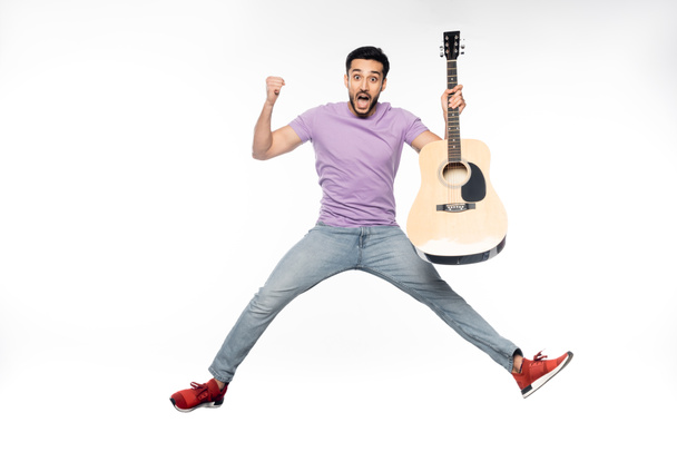uomo sorpreso in jeans blu e t-shirt viola levitante mentre tiene la chitarra acustica sul bianco  - Foto, immagini