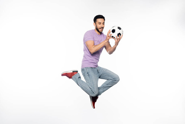счастливый человек в джинсах и фиолетовой футболке левитирует, глядя на футбол на белом  - Фото, изображение