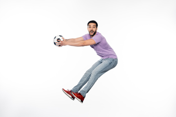 ενθουσιασμένοι άνθρωπος με τζιν και μωβ t-shirt αιωρείται με το ποδόσφαιρο σε λευκό  - Φωτογραφία, εικόνα