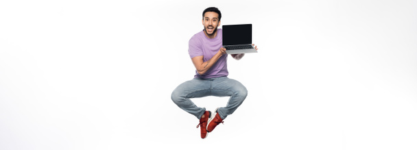 positive man in jeans and purple t-shirt levitating while holding laptop on white, banner - Photo, Image