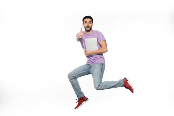 positive man in jeans and purple t-shirt levitating while holding laptop and showing thumb up on white  - Fotó, kép