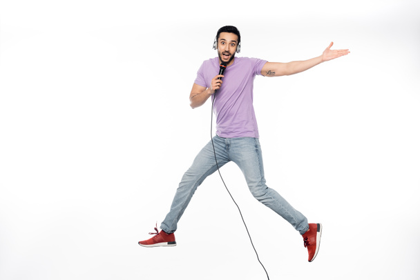 positive man in wireless headphones singing in microphone and levitating on white - Photo, Image