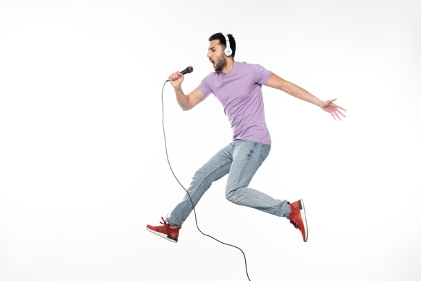 young man in wireless headphones singing in microphone and levitating on white - Photo, Image