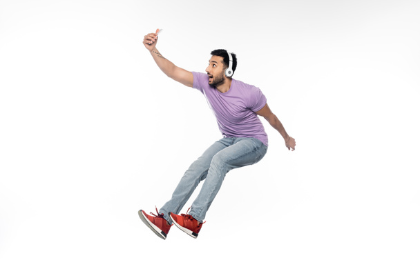 veselý muž v bezdrátových sluchátkách při selfie a levitaci na bílém - Fotografie, Obrázek