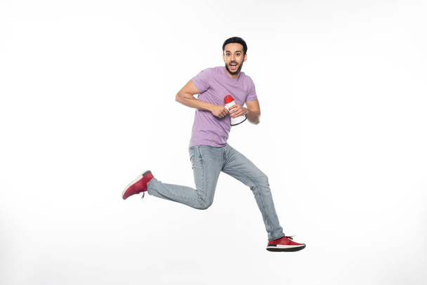 happy man in jeans and purple t-shirt levitating while holding megaphone on white  - Photo, Image