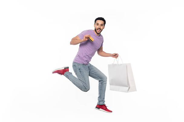 excited man in jeans and purple t-shirt levitating while holding credit card and shopping bag on white - 写真・画像
