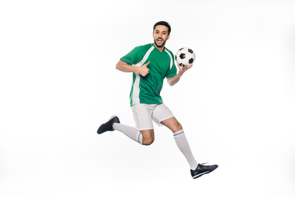 užaslý fotbalista v uniformě skákání při držení fotbalový míč a ukazující palec nahoru na bílém  - Fotografie, Obrázek