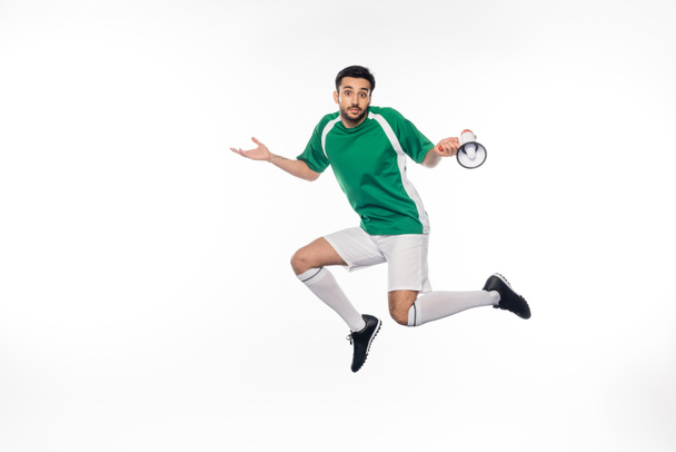 confused football player in uniform jumping and holding loudspeaker on white  - Fotoğraf, Görsel