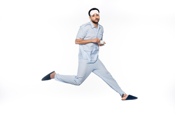 hombre barbudo en pijama sosteniendo taza y platillo mientras levita sobre blanco  - Foto, Imagen