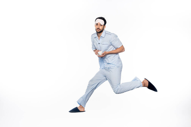 bearded man in pajamas nd sleeping mask holding cup with saucer while levitating on white  - Photo, image