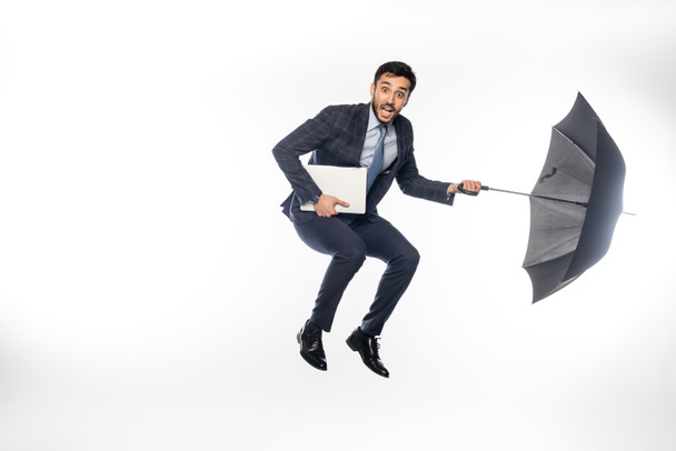 astonished businessman in suit jumping with laptop and umbrella on white  - Foto, imagen