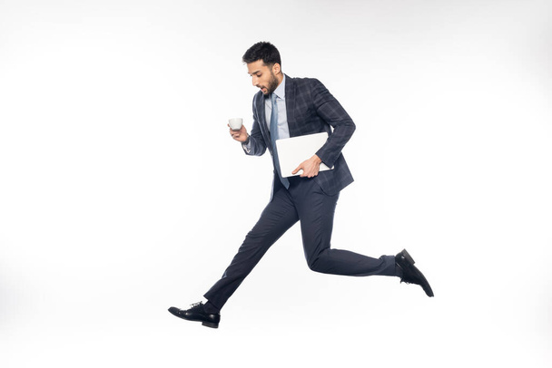 schockierter Geschäftsmann im Anzug springt, während er Laptop und Tasse auf weiß hält  - Foto, Bild