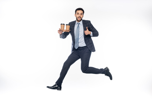 happy businessman in suit jumping while holding carton cup holder with paper cups and showing thumb up on white  - Foto, imagen
