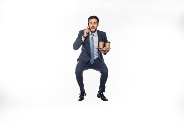 happy businessman in suit jumping while holding cup holder with paper cups and talking on smartphone on white  - 写真・画像