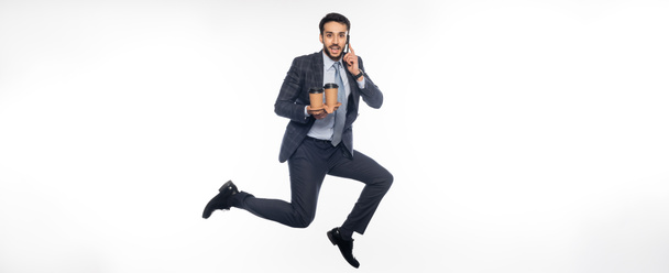 alegre hombre de negocios en traje saltando mientras sostiene el sostenedor de la taza con copas de papel y hablando en el teléfono inteligente en blanco, bandera - Foto, imagen