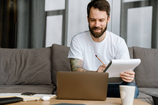 Bearded european man writing down notes while working with laptop at home - Foto, Imagem