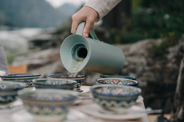 Hand pours a drink from a jug into cups on a background of mountains - Zdjęcie, obraz