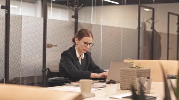 Cintura para arriba de la mujer de negocios caucásica de ojos marrones con discapacidades usando anteojos, sentada en la mesa en el espacio de coworking, usando computadora portátil - Metraje, vídeo