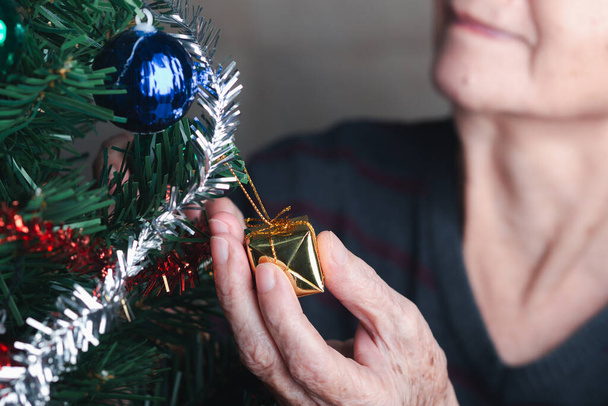 Close-up of hand senior woman decorating the Christmas tree while sitting in a living room at home. Space for text. Concept of aged people and Christmas and happy new year festival. - Fotó, kép