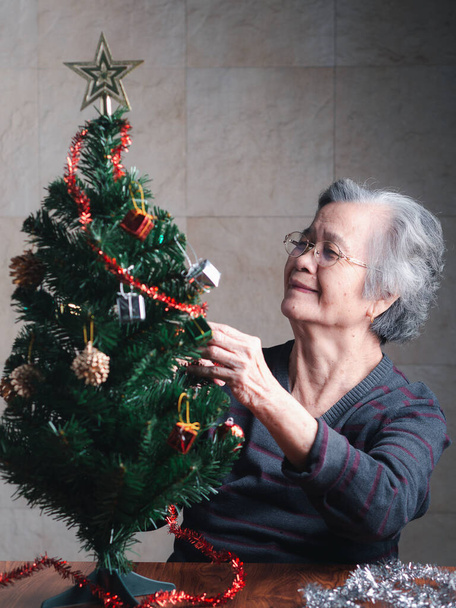 Porträt einer älteren Asiatin, die den Weihnachtsbaum schmückt, während sie zu Hause im Wohnzimmer sitzt. Raum für Text. Konzept für ältere Menschen und Weihnachten und ein frohes neues Jahr. - Foto, Bild