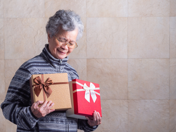 Portrait of an elderly Asian woman hug gift boxes, smiling and looking down while standing in a living room at home. Space for text. Concept of aged people and Christmas and happy new year festival. - Foto, Imagem