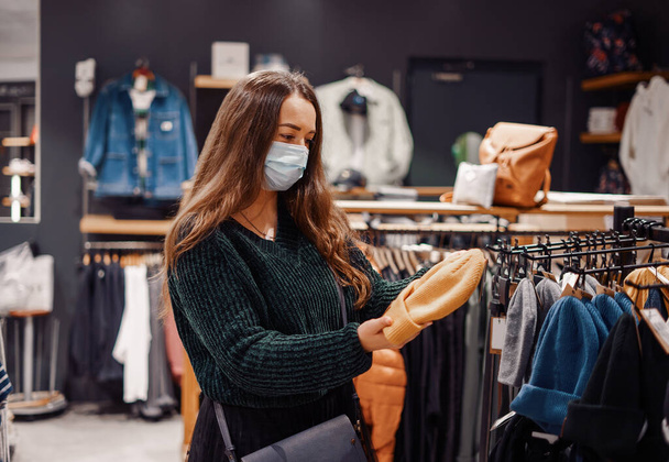 Mujer joven con pelo largo suelto y mascarilla haciendo compras de temporada de ropa - Foto, imagen