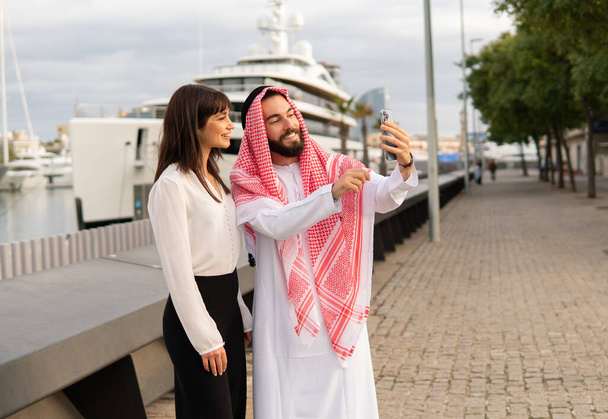 Усміхнений арабський бізнесмен бере селфі зі своїм молодим привабливим європейським жіночим помічником у порту
 - Фото, зображення