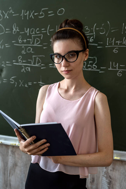 若い深刻な女性教師は、黒板の横に大学で数式を説明します   - 写真・画像