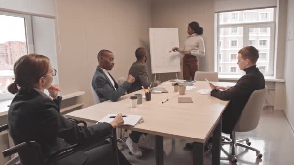 Steadicam of young woman presenting business graph to African American and Caucasian men and female colleague in wheelchair sitting at table in conference room at daytime - Footage, Video