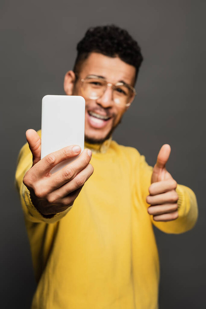 homme afro-américain flou et positif dans des lunettes prenant selfie isolé sur gris - Photo, image
