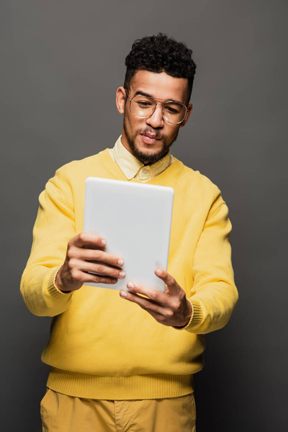 Genç Afro-Amerikan gözlüklü adam gri üzerinde izole edilmiş dijital tablet kullanıyor. - Fotoğraf, Görsel