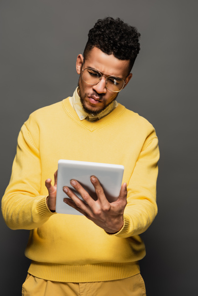 joven afroamericano hombre en gafas usando borrosa tableta digital aislado en gris - Foto, Imagen