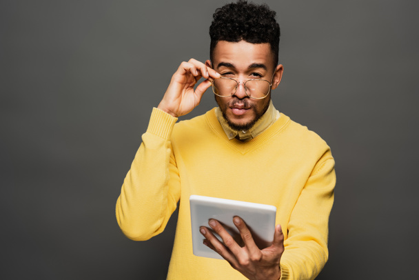 focused african american man adjusting glasses and holding digital tablet on dark grey - 写真・画像
