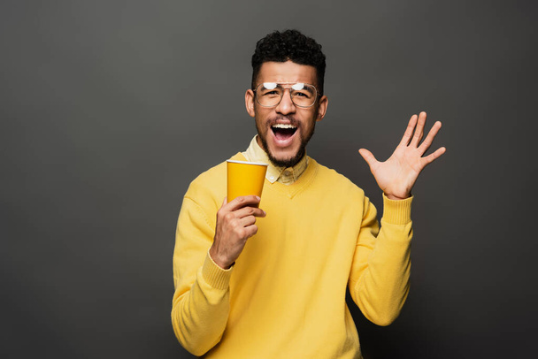 excité homme afro-américain dans des lunettes tenant tasse en papier et geste sur gris foncé - Photo, image